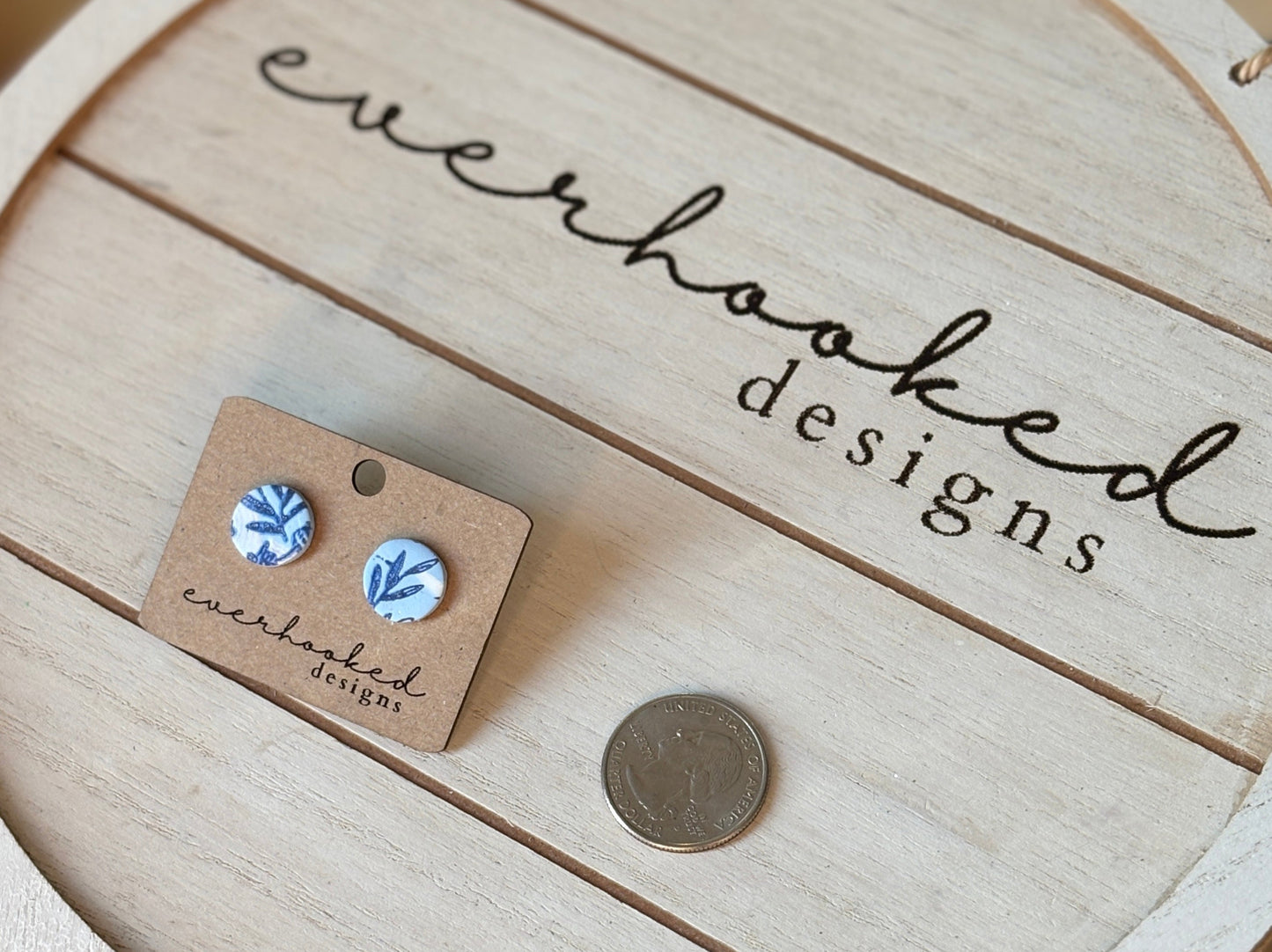 Clay blue stud earrings featuring a painted blue floral design Placed next to a quarter for sizing