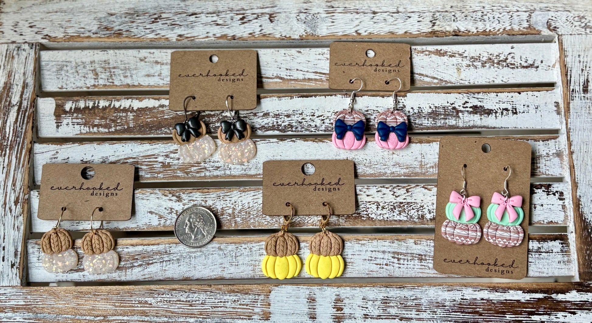 Display of polymer clay double pumpkin dangly earrings (with and without bows) on a rustic wooden board. Shown with a quarter for sizing.