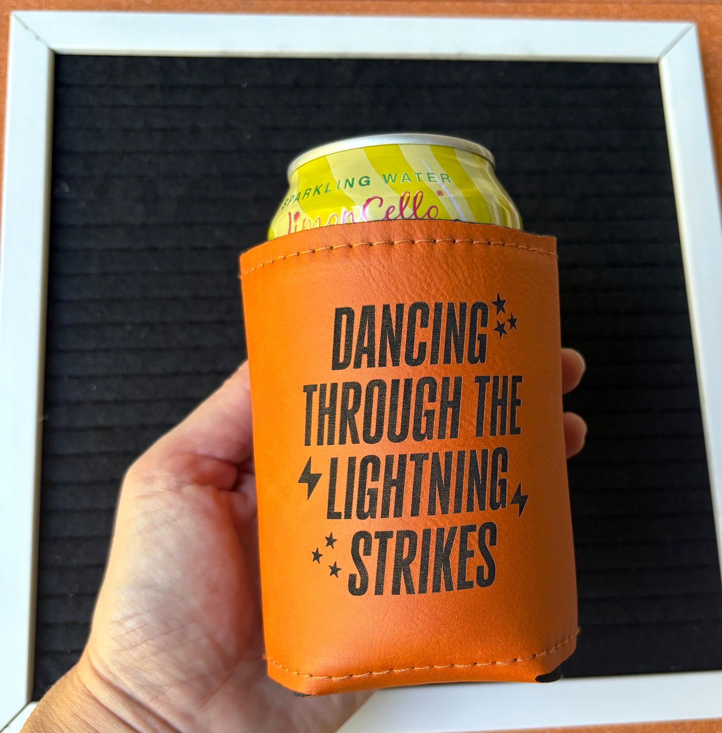 Orange leatherette koozie with text "Dancing Through the Lightning Strikes" held by a hand against a textured surface