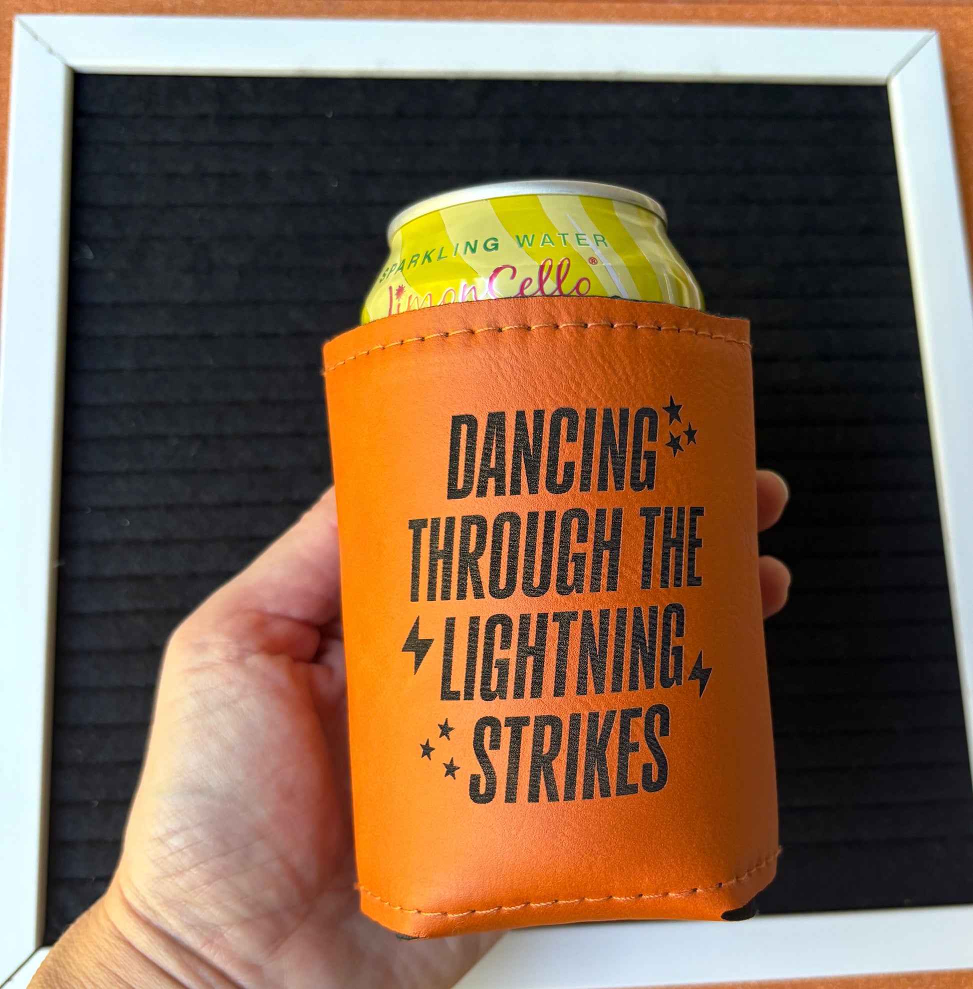 Orange leatherette koozie with text "Dancing Through the Lightning Strikes" held by a hand against a textured surface