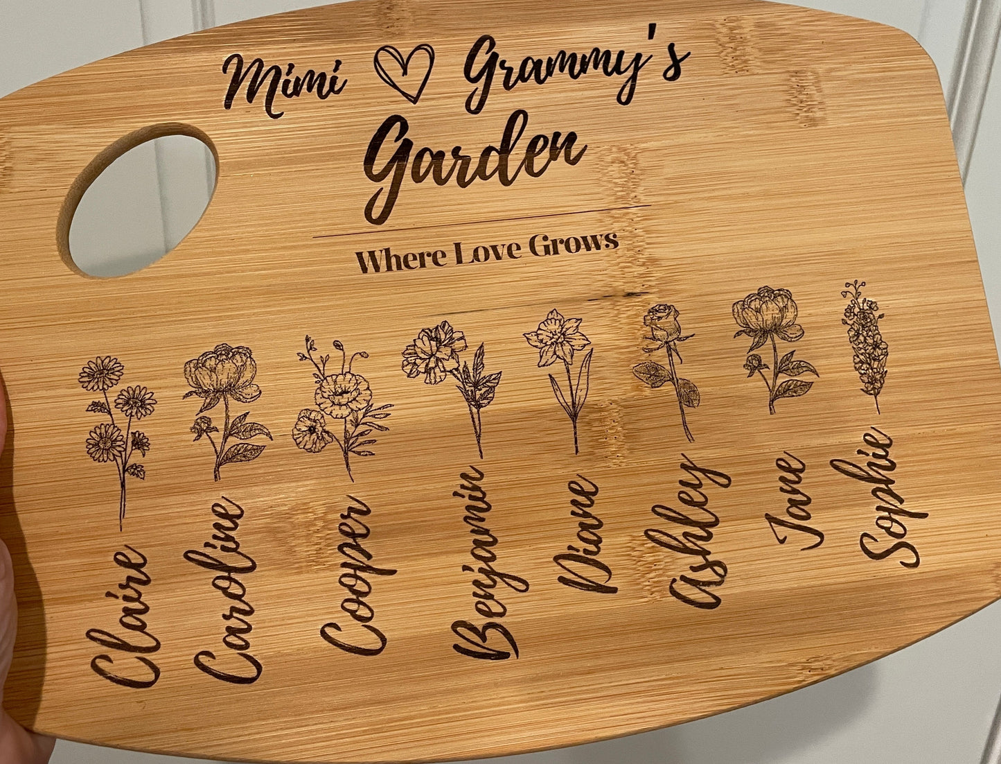Cutting board with Mimi, a heart, Grammy's Garden on top, then "Where Love Grows," and then below that eight names with corresponding birth flowers.