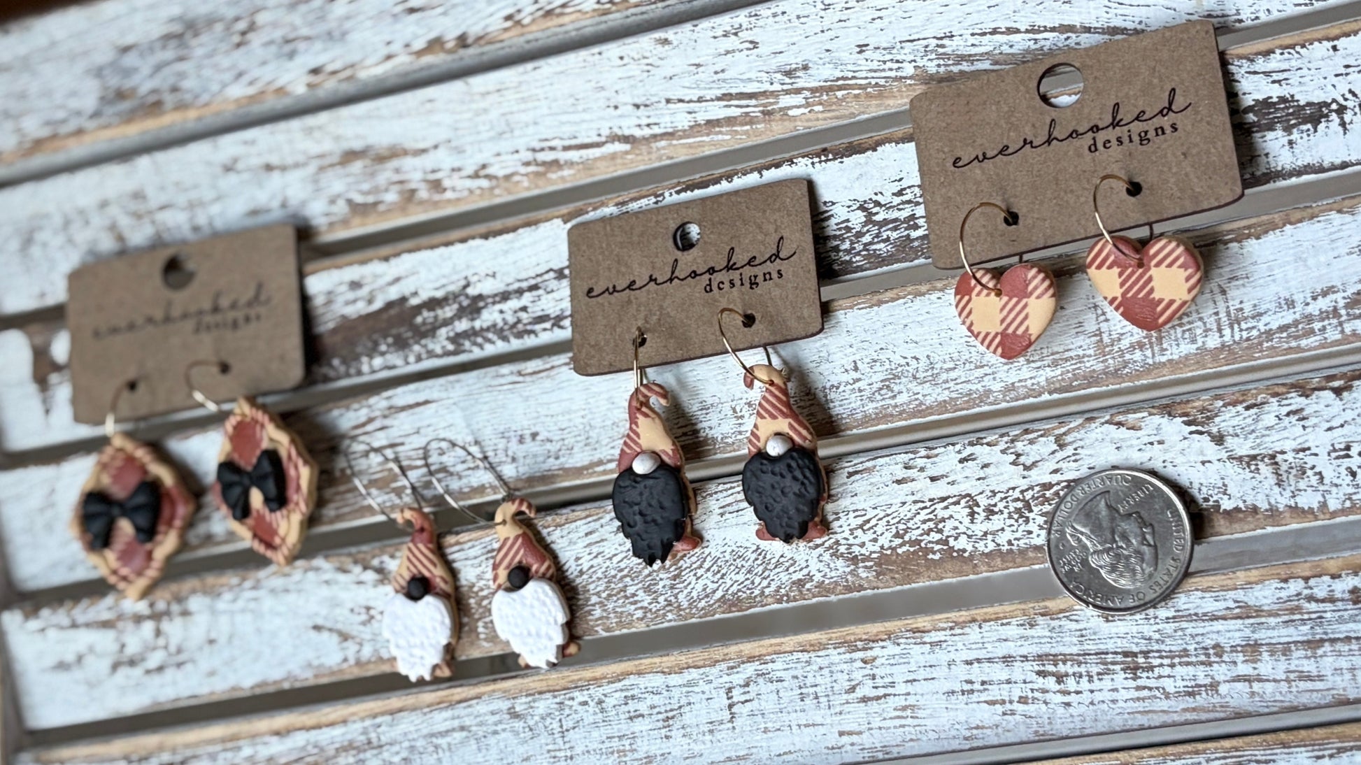 Handmade polymer clay earrings shaped like framed ribbon, whimsical gnomes and hearts on hoops. Each pair has a red and beige plaid design, lightweight and seasonal. Placed next to a quarter for sizing.