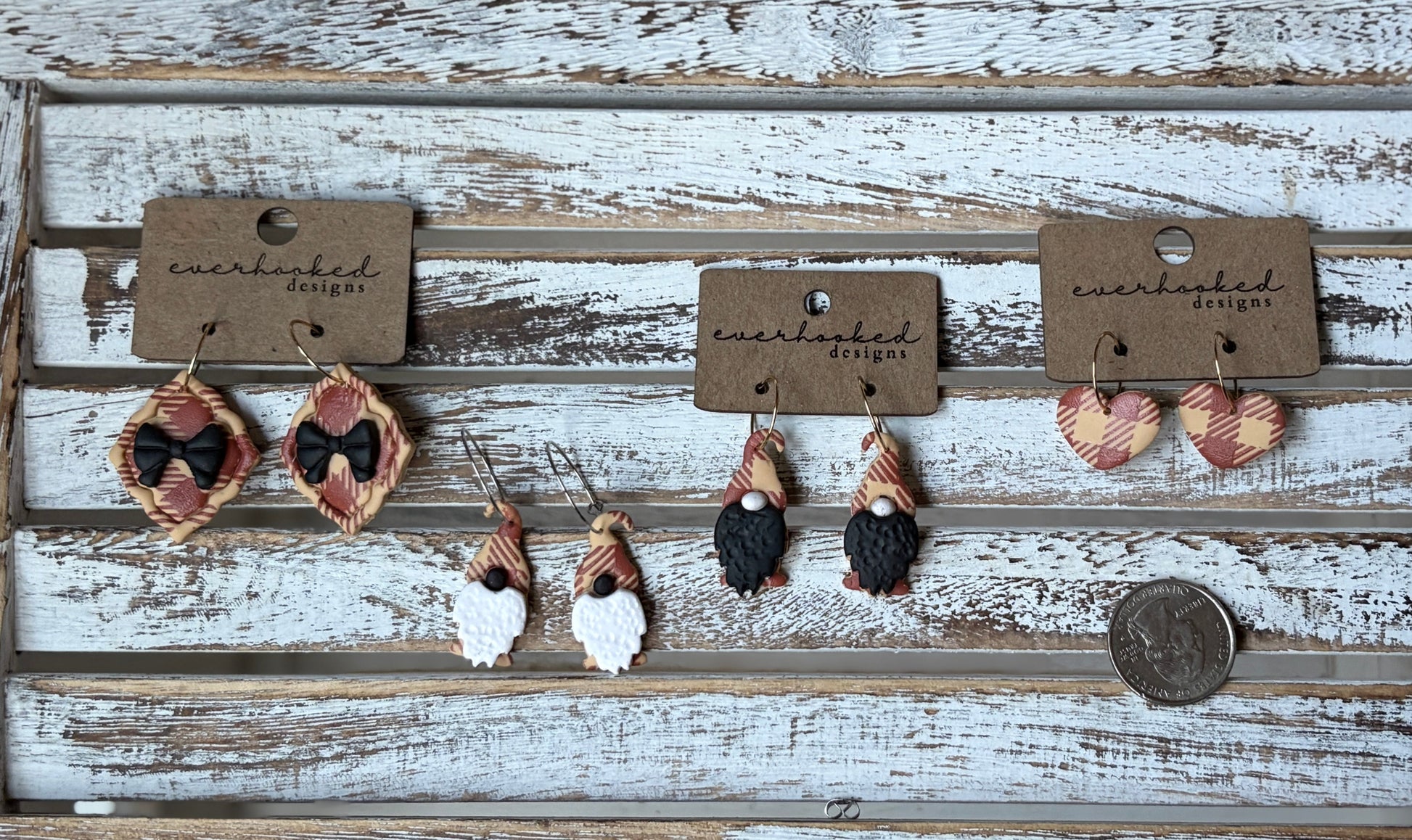 Handmade polymer clay earrings shaped like framed ribbon, whimsical gnomes and hearts on hoops. Each pair has a red and beige plaid design, lightweight and seasonal. Placed next to a quarter for sizing