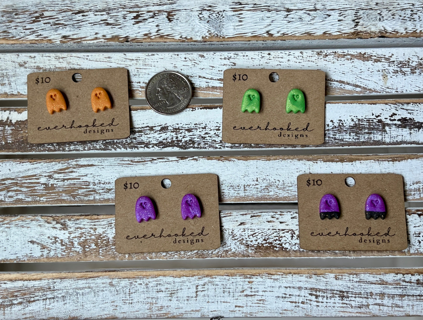 Handmade polymer clay ghost earrings in green, orange, and purple displayed on rustic wood background. Pictured with a quarter for sizing.