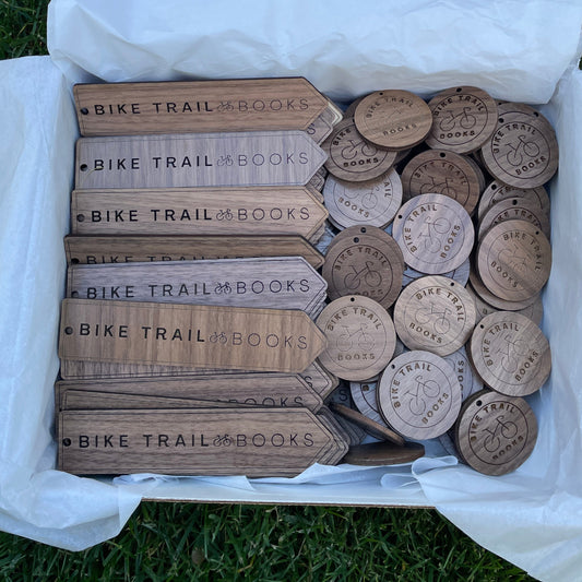 Box of custom laser-engraved wooden bookmarks and round keychains for Bike Trail Books, displayed on white tissue paper over grass