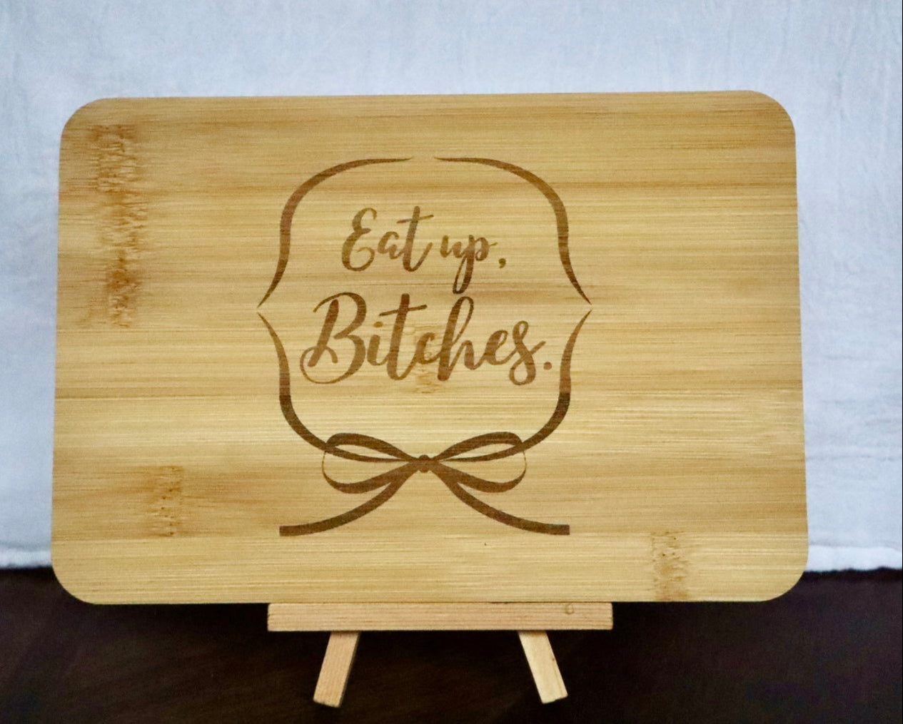 "Eat up, Bitches" with a ribbon around it on a bamboo cutting board