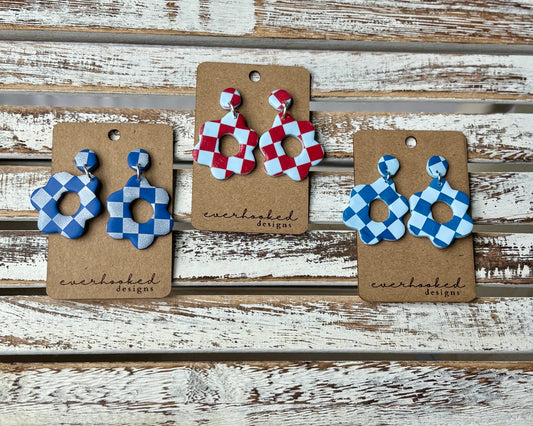 Three pairs of checkerboard clay earrings in a floral cutout shape, displayed on kraft earring cards. Color combos include red and light blue, navy and light blue, and bright blue and white.