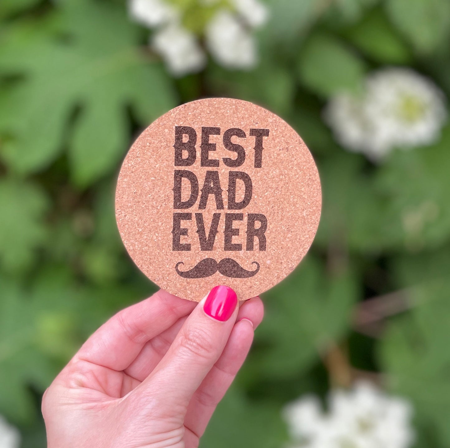 Cork coaster engraved with the phrase “BEST DAD EVER” and a mustache design, held in front of greenery and white flowers.