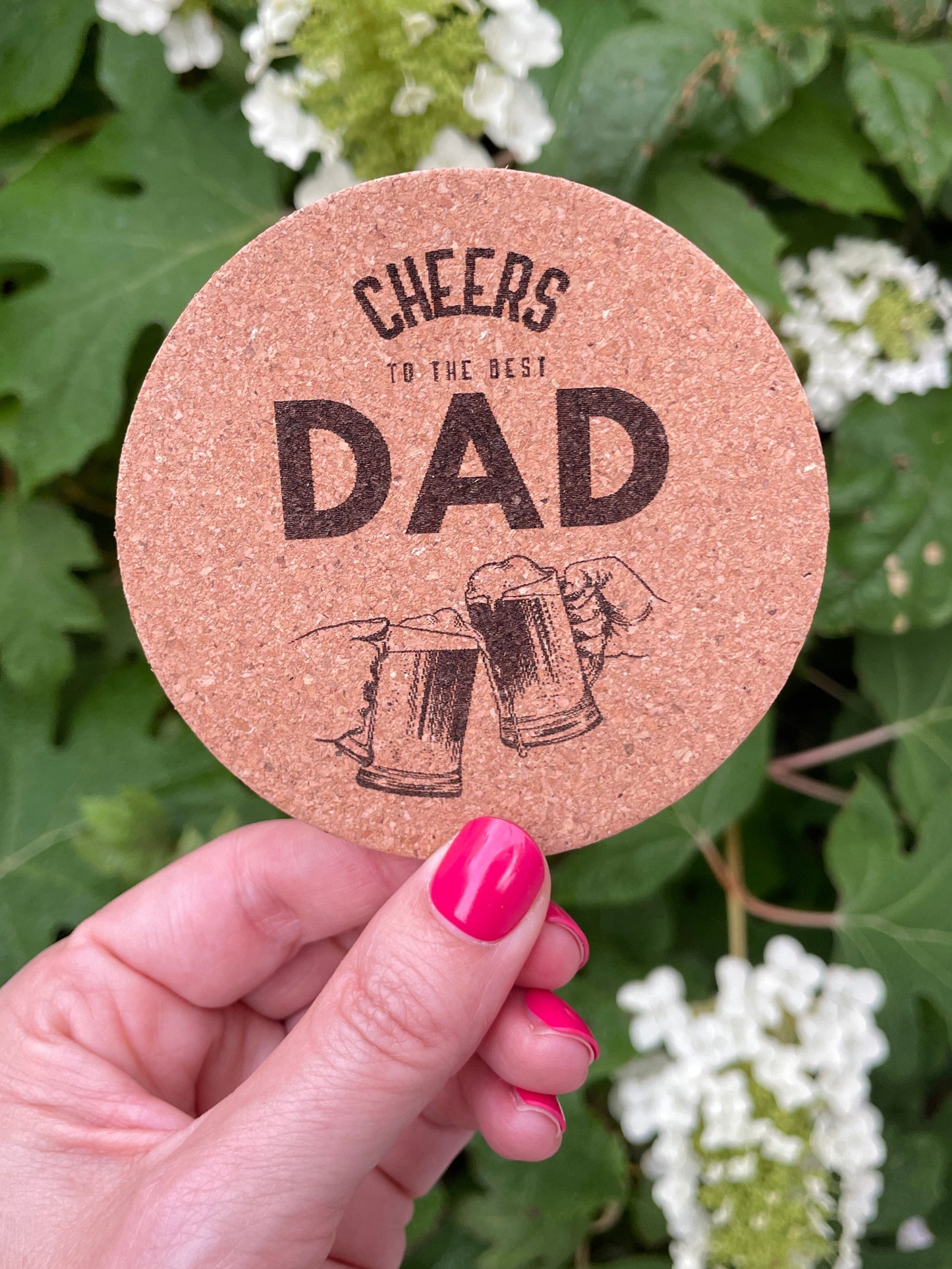 Cork coaster engraved with the phrase “CHEERS TO THE BEST DAD” and two clinking beer mugs, held in front of leafy greenery and white flowers.
