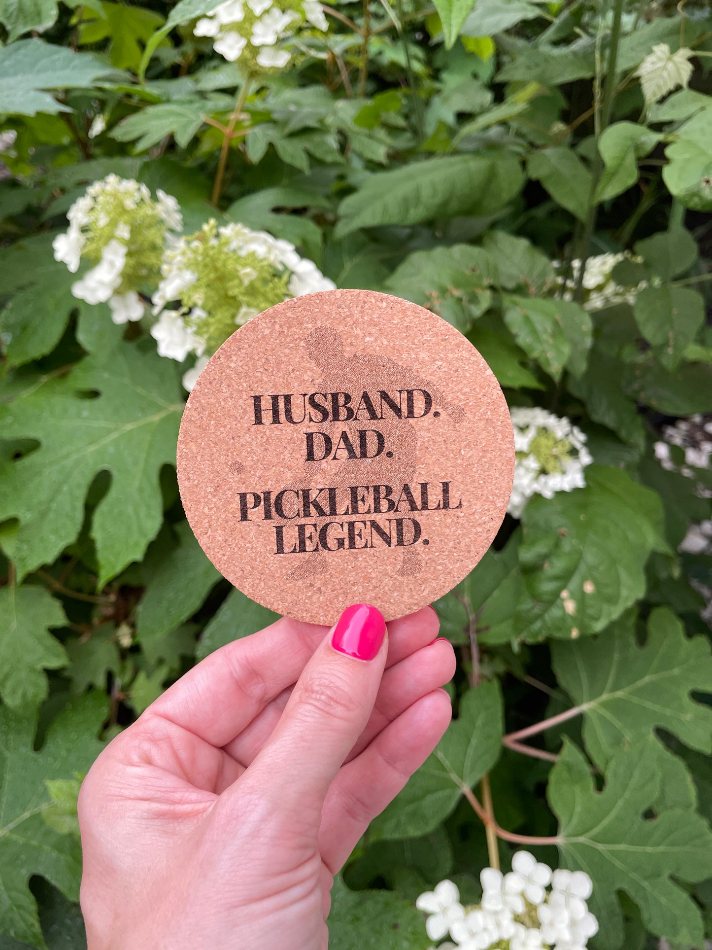 Engraved cork coaster with the phrase “HUSBAND. DAD. PICKLEBALL LEGEND.” held in front of white hydrangea flowers and green leaves.