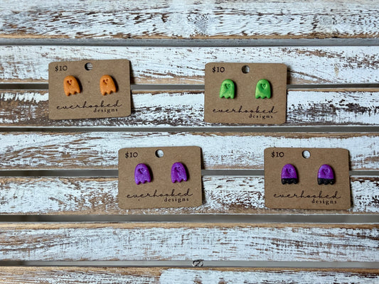 Handmade polymer clay ghost earrings in green, orange, and purple displayed on rustic wood background