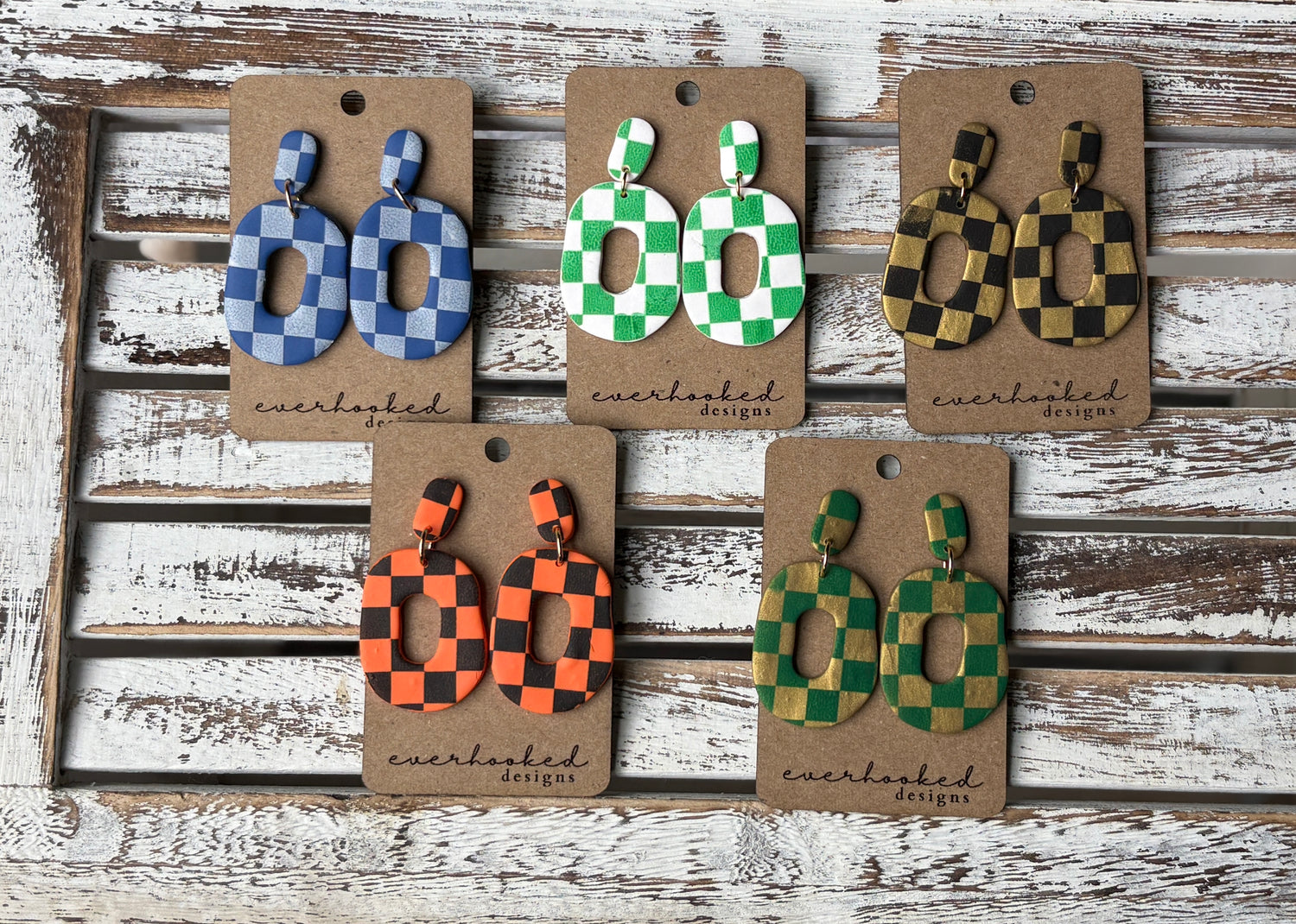 Assorted handmade polymer clay earrings in a checkerboard pattern, displayed in various shapes and bold color combinations including blue, green, orange, black, gold, and white