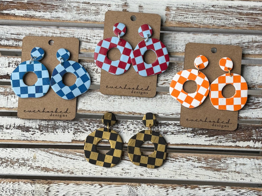 Assorted handmade polymer clay earrings in a checkerboard pattern, displayed in wavy circle designs and bold color combinations including red, blue, green, orange, black, gold, and white