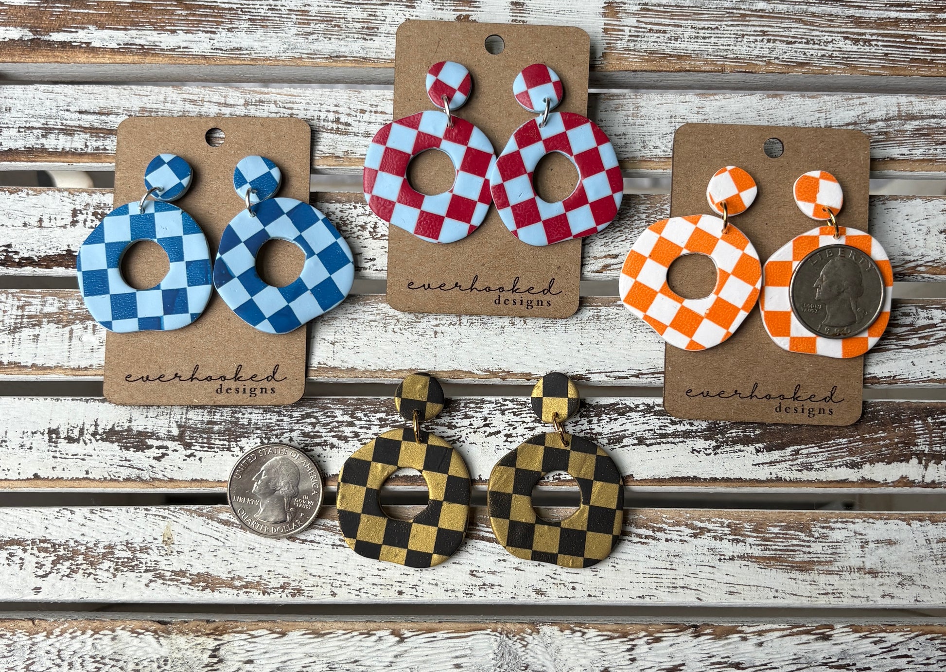 Assorted handmade polymer clay earrings in a checkerboard pattern, displayed in wavy circle designs and bold color combinations including red, blue, green, orange, black, gold, and white. Featured with quarters to demonstrate size.