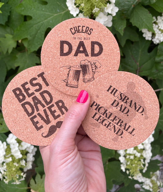 Three engraved cork coasters with Father’s Day messages: “BEST DAD EVER,” “CHEERS TO THE BEST DAD,” and “HUSBAND. DAD. PICKLEBALL LEGEND.” held in one hand in front of blooming hydrangeas.